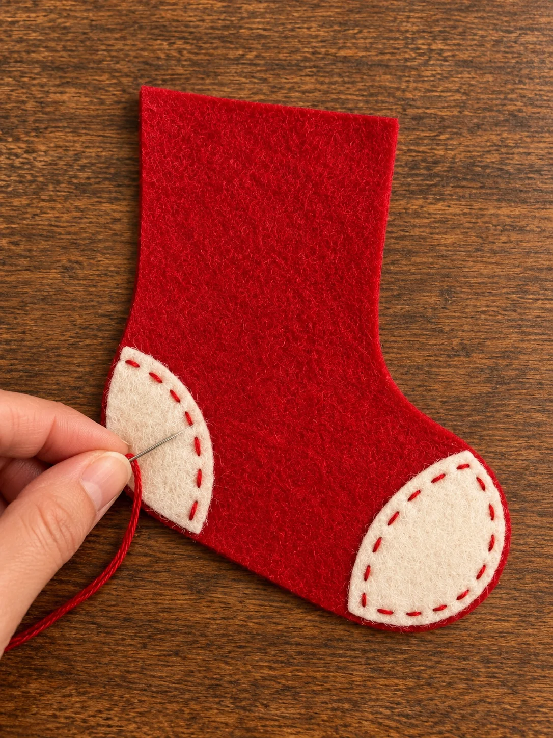 One hand stitching an ivory heel patch to a red felt stocking on warm dark wood