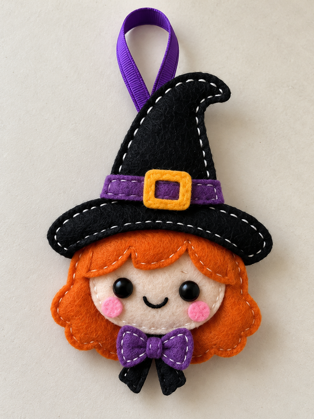 Illustrative felt witch ornament with orange hair, a curled black hat, yellow buckle, purple bow, and hanging loop