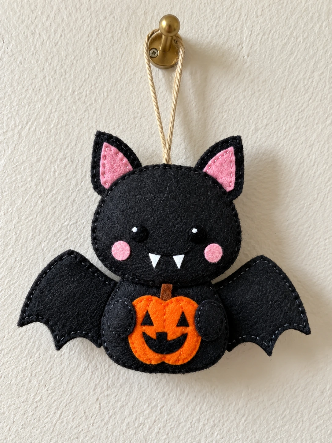 Black felt pumpkin bat ornament hanging from twine on a wall hook, holding an orange jack-o’-lantern