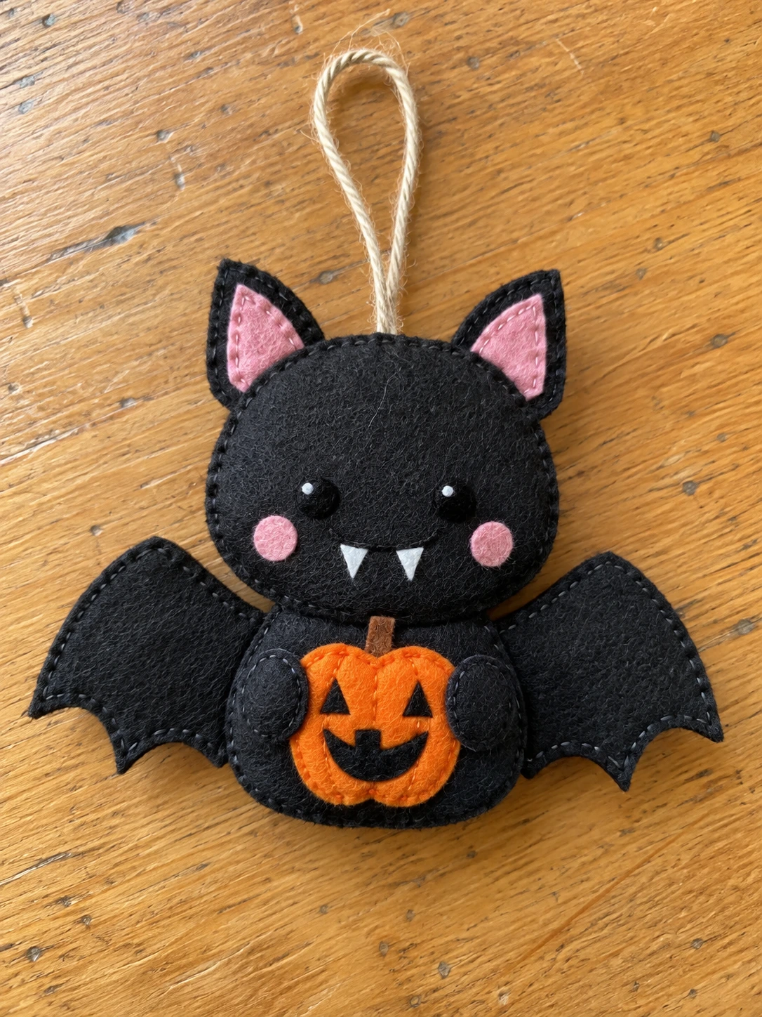 Black felt pumpkin bat ornament on a wooden table, with pink ears and cheeks, white fangs, and little paws holding an orange jack-o’-lantern