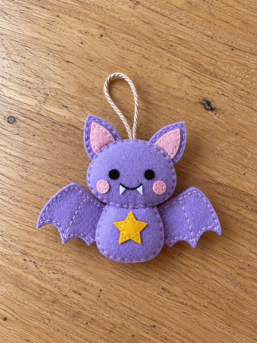 Lavender felt star bat ornament on a wooden table, with pink ears and cheeks, black eyes, white fangs, and a golden star